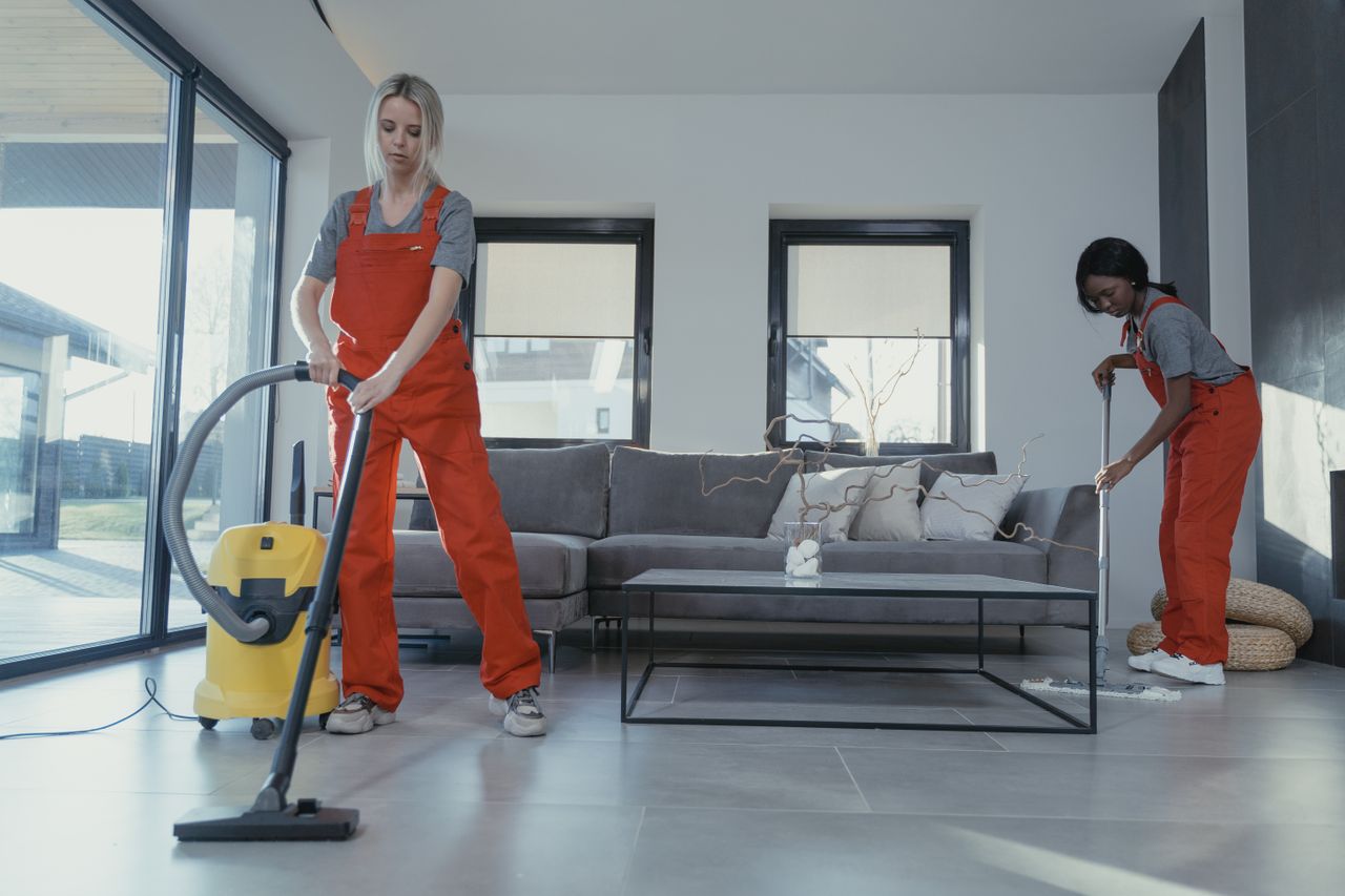 services-05 Two women in red overalls vacuuming and sweeping a modern living room.